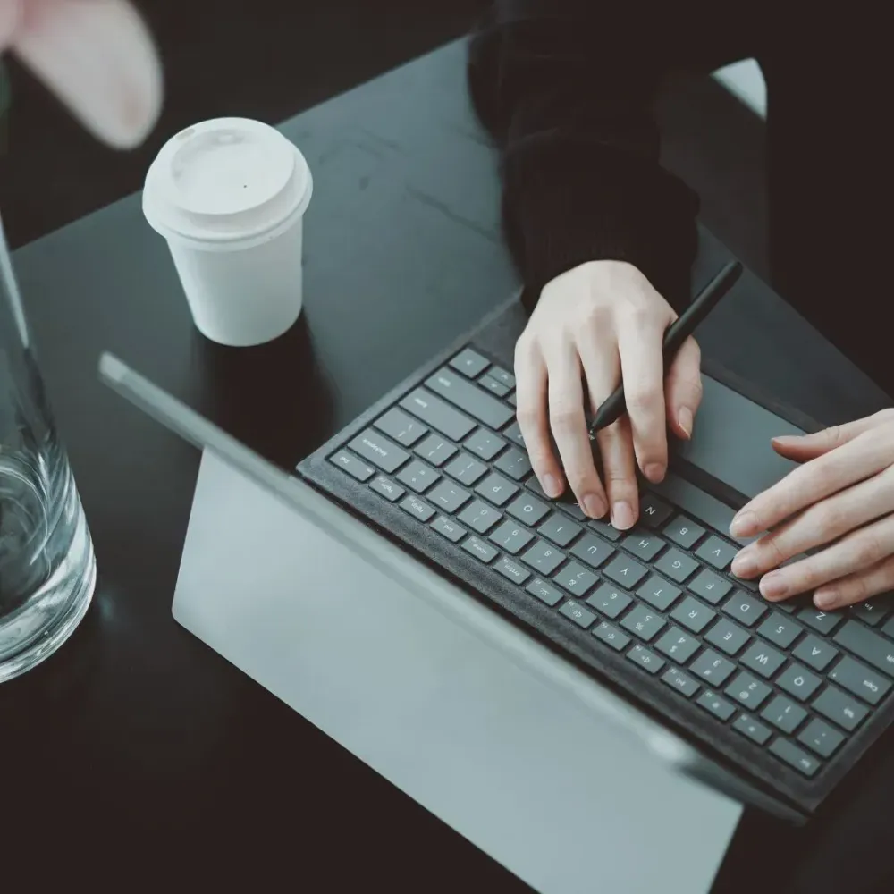 Eine Person die in einem Tisch am Laptop arbeitet und einen Becher Kaffee rechts neben sich stehen hat